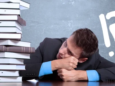 A law student in a suit rests his head beside a tall stack of books, with question and exclamation marks on a chalkboard behind him.