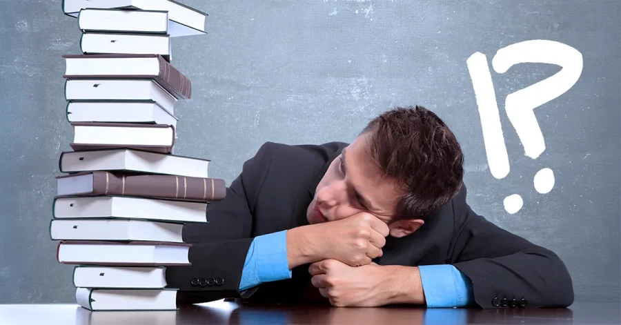 A law student in a suit rests his head beside a tall stack of books, with question and exclamation marks on a chalkboard behind him.