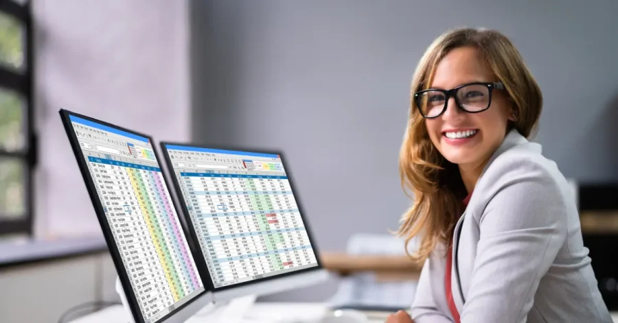 A smiling working woman wearing glasses and a light blazer sits at a desk with two monitors in front of her in a modern office.