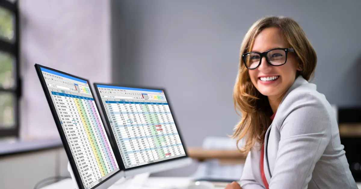 A smiling working woman wearing glasses and a light blazer sits at a desk with two monitors in front of her in a modern office.