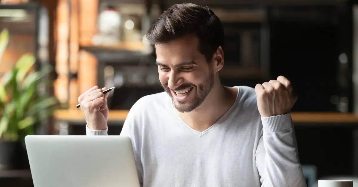 Law student sitting at a desk, celebrating with clenched fists while looking at a laptop screen, holding a pen in one hand.