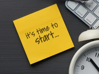 Yellow sticky note with handwritten text “It’s time to start...” placed on a dark desk surface next to a white alarm clock and a partial view of a computer keyboard, symbolizing motivation and productivity.