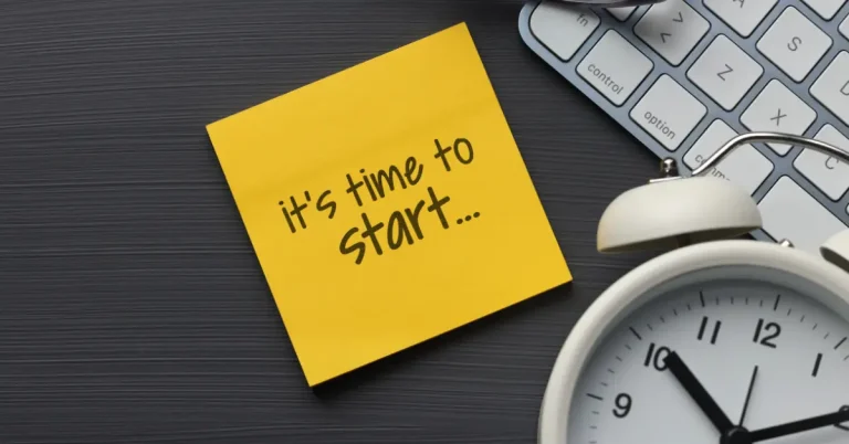 Yellow sticky note with handwritten text “It’s time to start...” placed on a dark desk surface next to a white alarm clock and a partial view of a computer keyboard, symbolizing motivation and productivity.