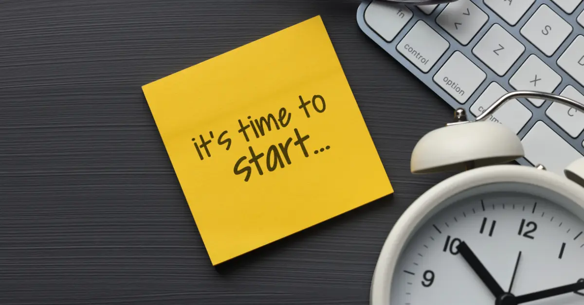Yellow sticky note with handwritten text “It’s time to start...” placed on a dark desk surface next to a white alarm clock and a partial view of a computer keyboard, symbolizing motivation and productivity.