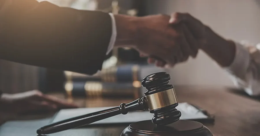 Close-up of a judge’s gavel resting on a legal document, with two individuals in business attire shaking hands in the background, symbolizing a job offer.