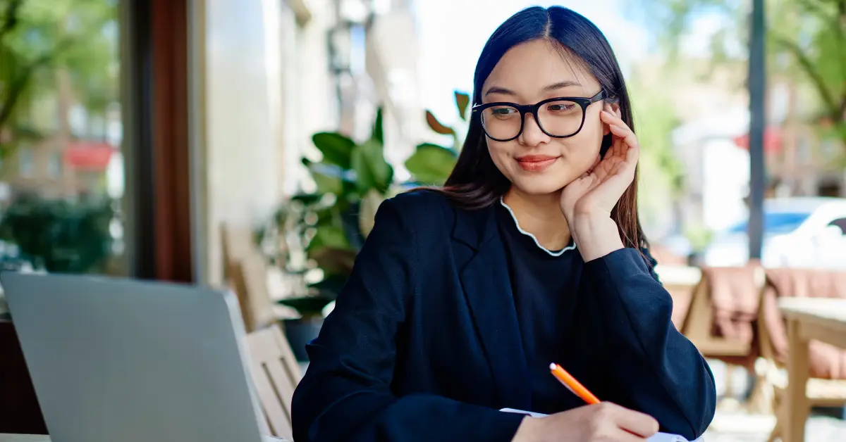 Woman in glasses and a black blazer sitting at an outdoor café, looking at a laptop and writing in a notebook.
