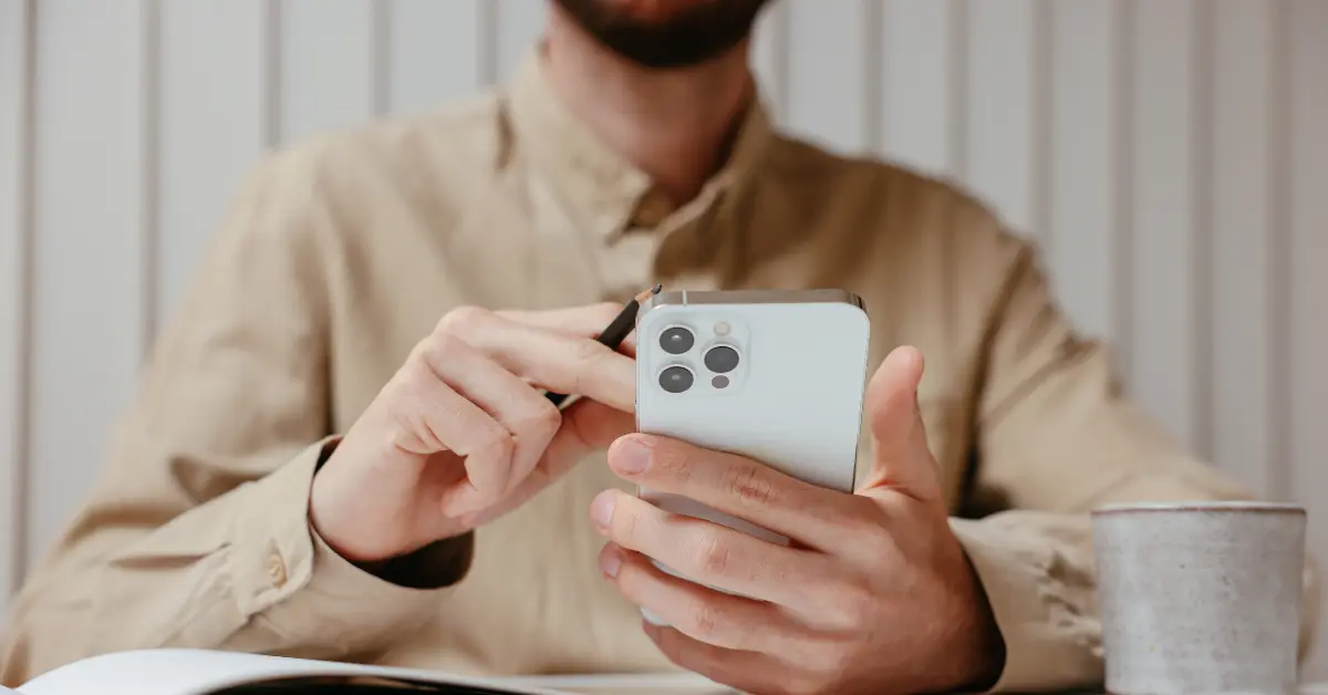 Student holding a smartphone with a pencil, wearing a beige shirt, seated at a desk with a cup and an open notebook.