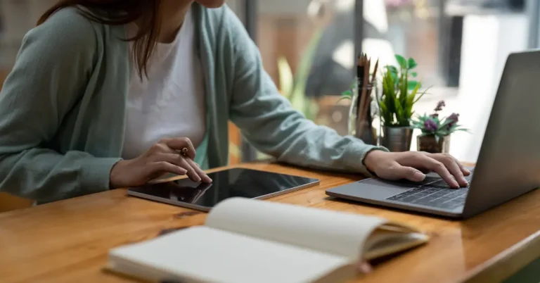 Woman working at a desk using both a tablet and laptop, with an open notebook and potted plants nearby, in a well-lit room with large windows.