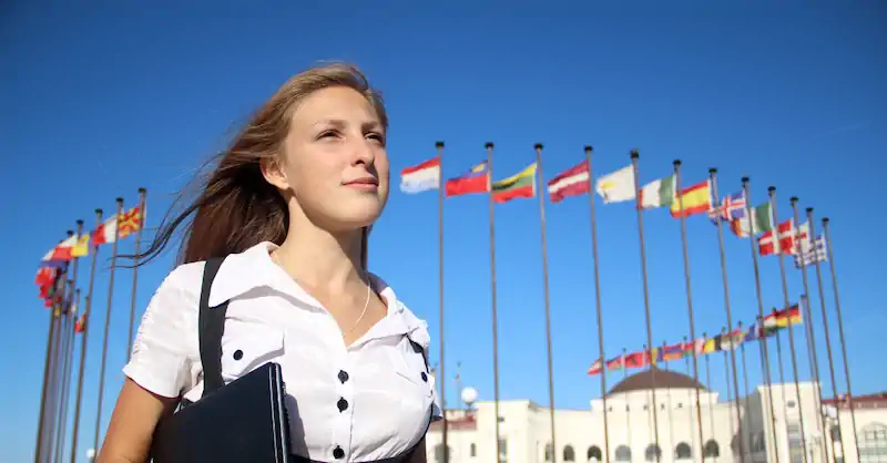 Confident young woman standing in front of a row of international flags against a clear blue sky, suggesting a global or diplomatic setting.