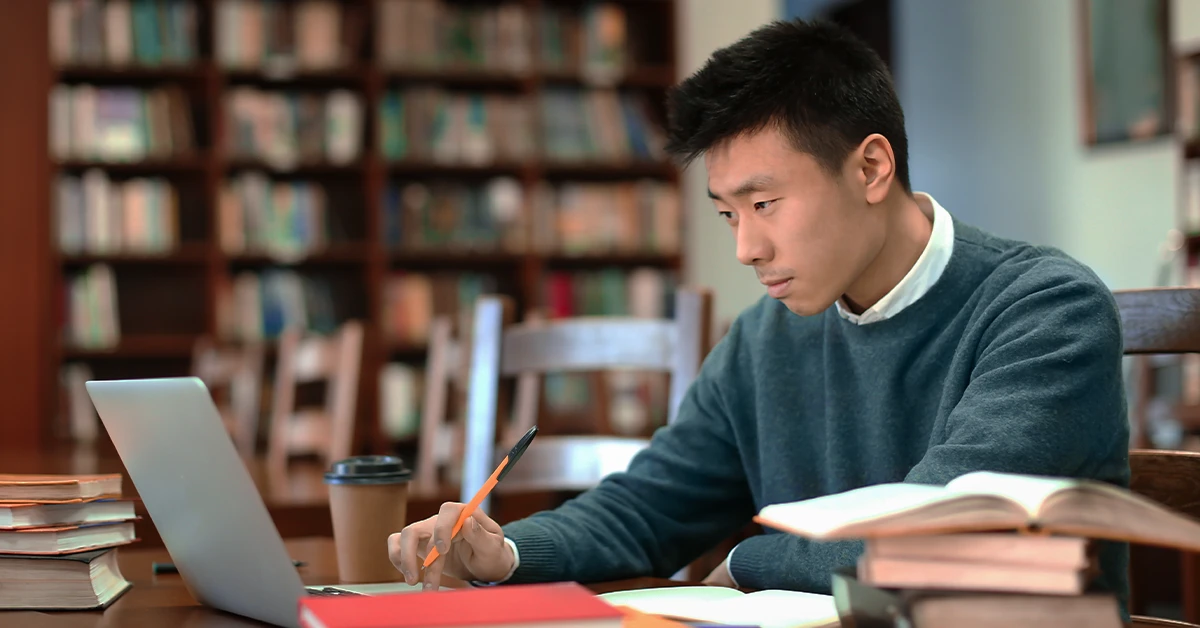 Focused student studying for the bar exam in a library with a laptop, open books, and coffee, researching the best bar exam prep courses for 2025.